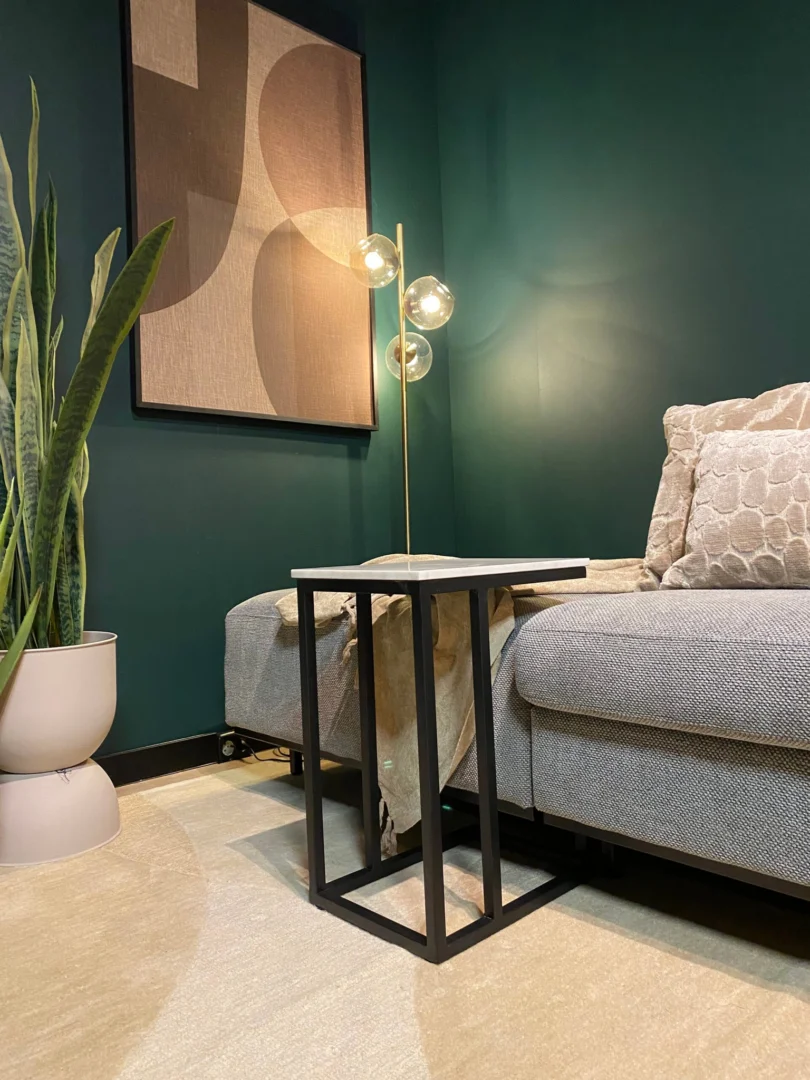 Een moderne woonkamer heeft donkergroene muren, een beige bank met gedessineerde kussens, abstracte muurkunst, een gouden vloerlamp met ronde lampen, een hoge plant, een minimalistische zwart metalen bijzettafel en de Schuifregelaar Nova Wit marmer.