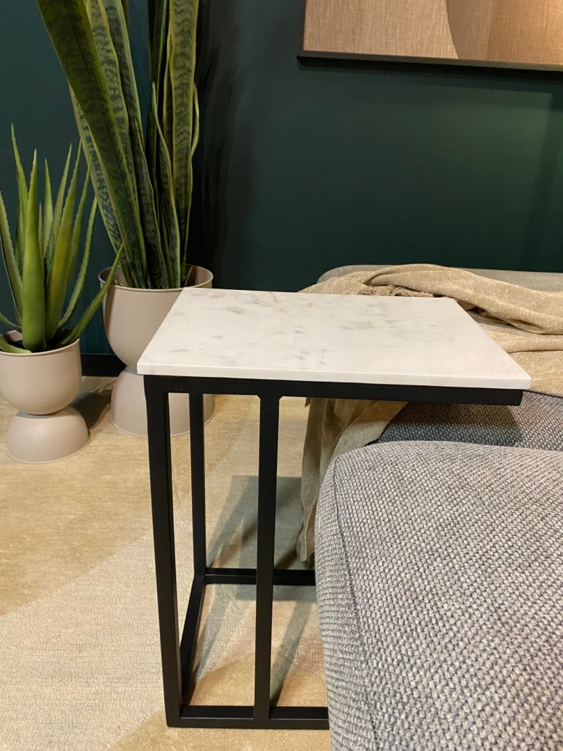 De Schuifregelaar Nova Wit marmer, een compacte bijzettafel met een wit marmeren blad en een zwart metalen onderstel, staat naast een grijs gestoffeerde bank. Op de achtergrond staan twee groene potplanten op een beige vloerkleed bij een donkergroene muur.