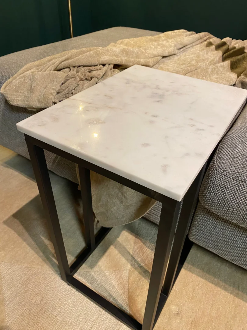 De Schuifregelaar Nova Wit marmer, een klein bijzettafeltje met een wit marmeren blad en zwarte metalen poten, staat naast een grijze bank gedrapeerd met een beige structuurdeken voor een gezellige, moderne uitstraling.