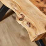 Close-up van de Boomstamtafel Live Edge 240 cm, met een naturel houten tafelblad met een ongelijke rand, zichtbare nerven en knoesten, en een klein gat. De tafel rust op donkere metalen poten op een lichte hardhouten vloer.