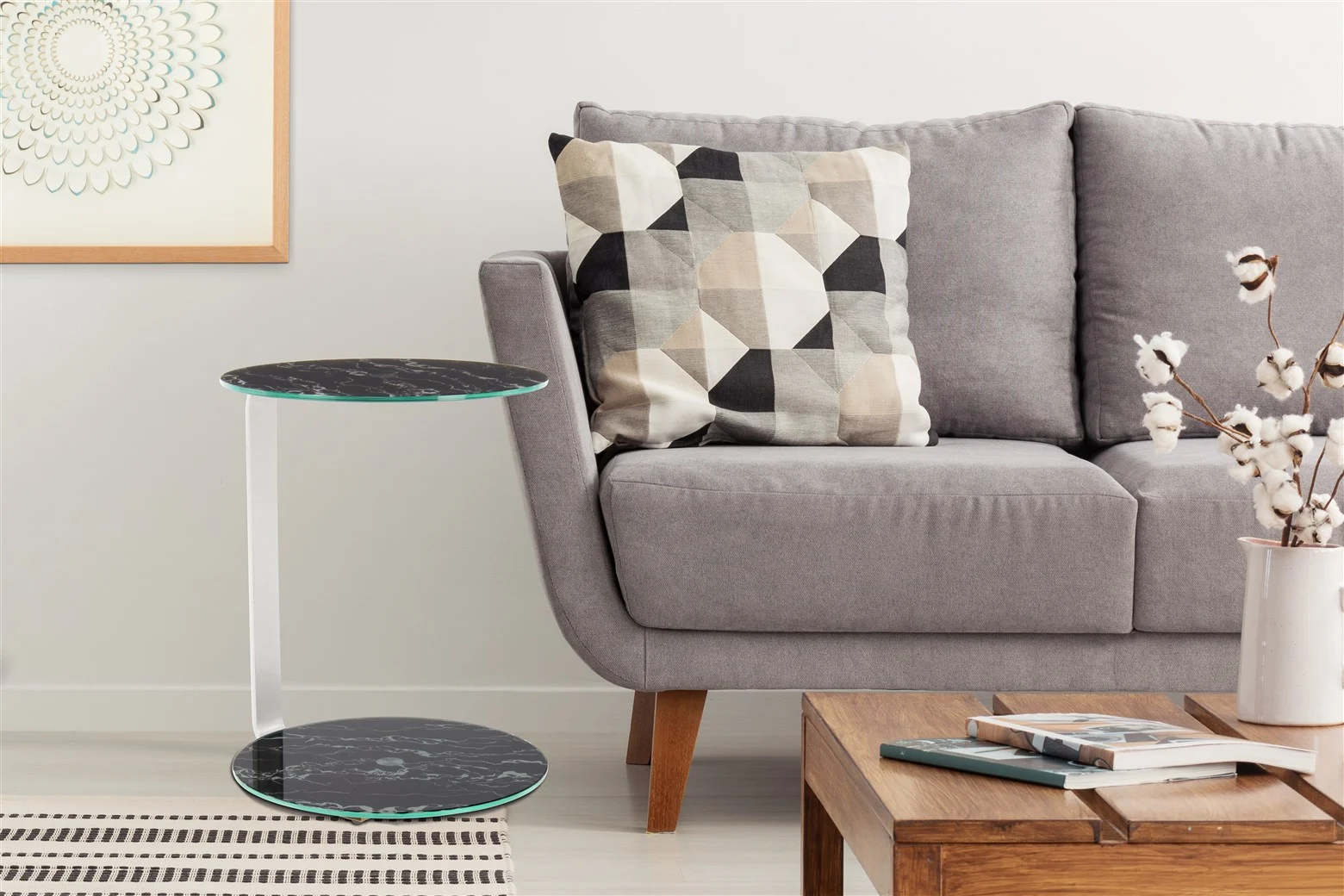 Een moderne woonkamer met een grijze bank, een geometrisch kussen, een compacte zwarte Bijzettafel Zilver, een houten salontafel met boeken, een vaas met katoenen stengels en een ingelijst abstract kunstwerk aan de muur.