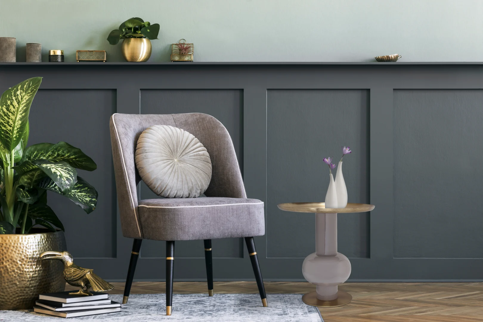 Een moderne woonkamer met een grijze accentmuur, een grijs gestoffeerde stoel met een rond kussen, de Bijzettafel Taupe, Goud Ijzer als bijzettafel, een witte vaas, gouden decoratieve items en groene planten in gouden potten.