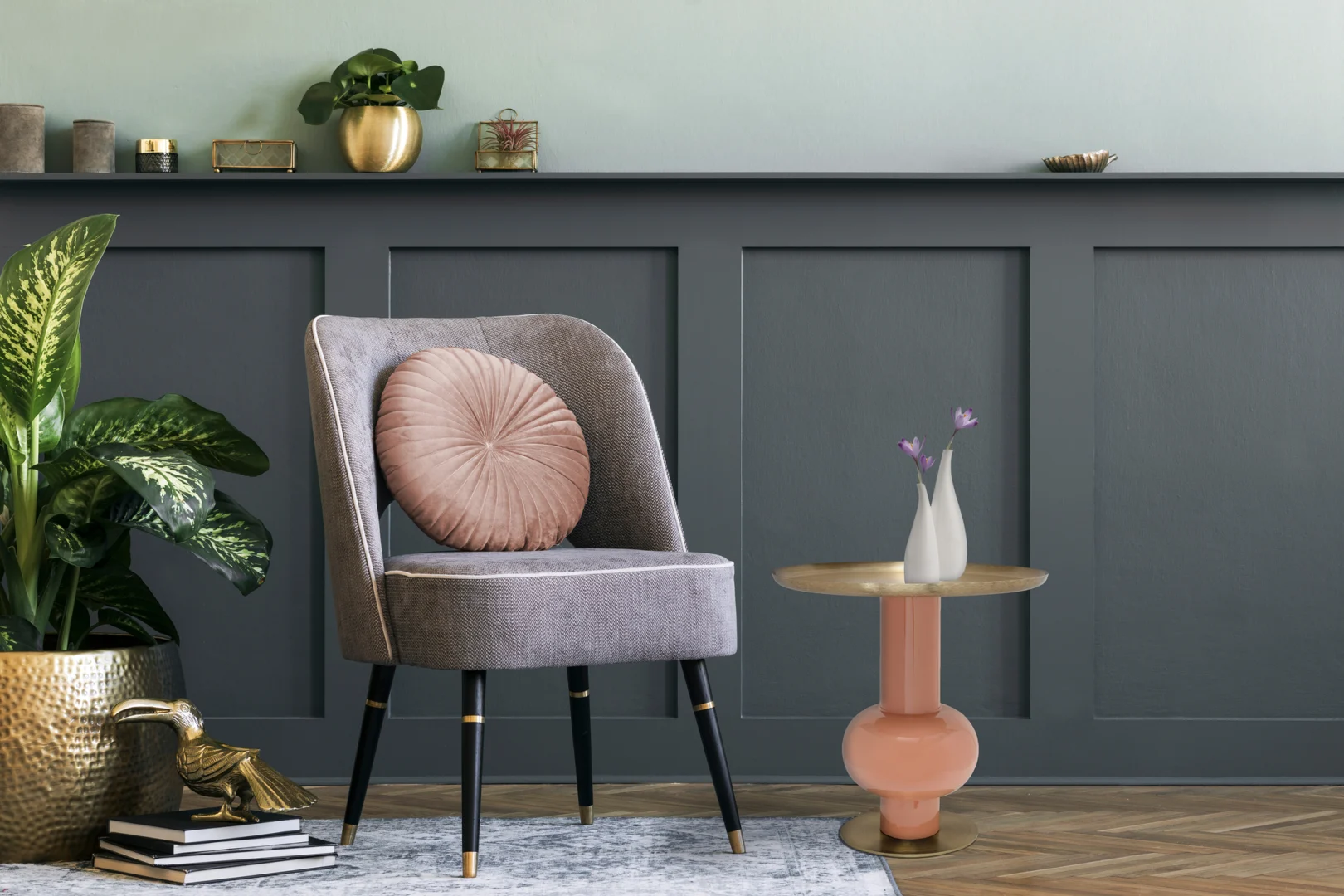 Een moderne woonkamer met een grijs gestoffeerde stoel, een rond roze kussen, de Bijzettafel Goud Ijzer met een witte vaas met paarse bloemen, groene planten en decoratieve accenten tegen een grijze lambrisering.