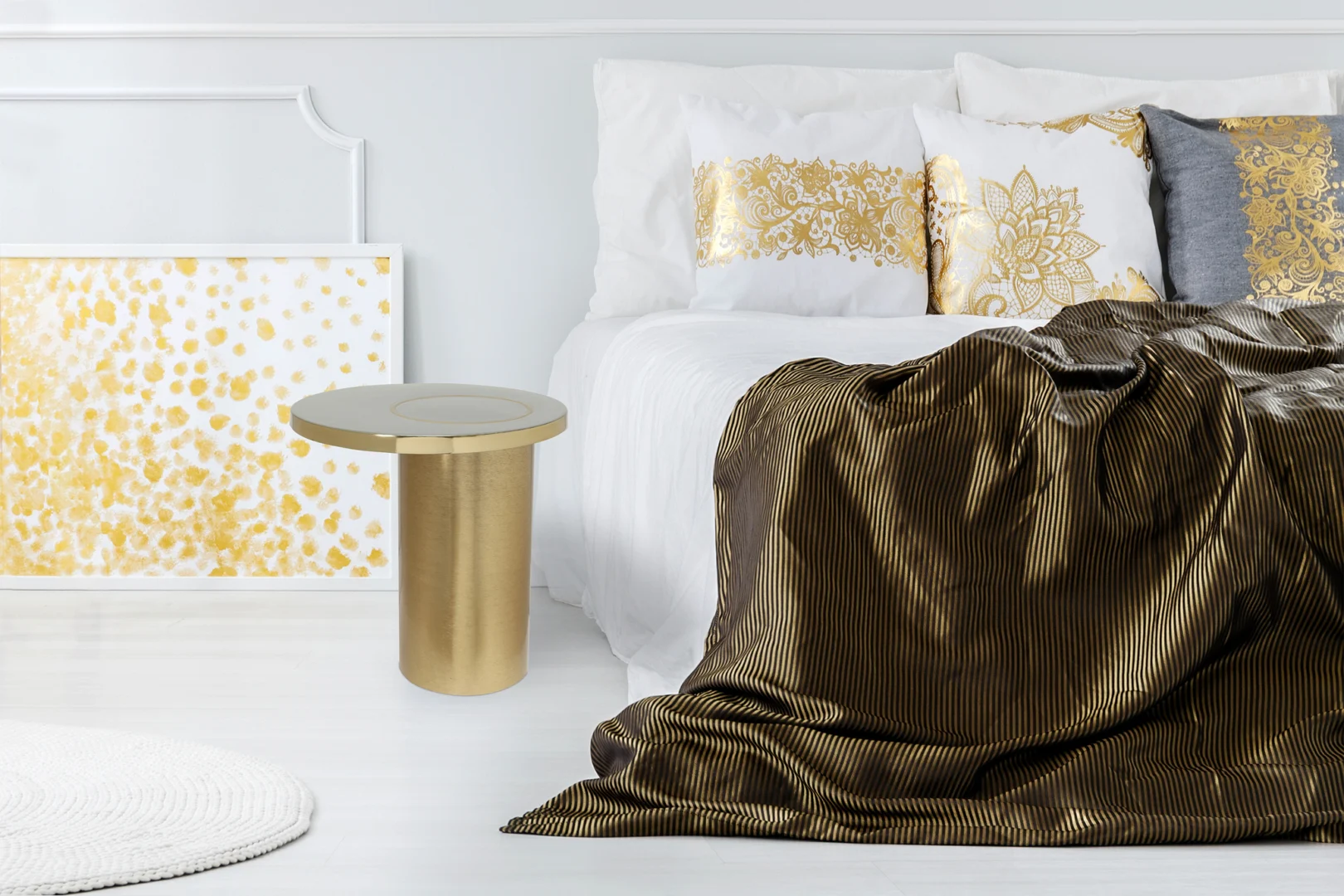 Een moderne slaapkamer heeft een sprei met goudstrepen, witte en gouden kussens, een Bijzettafel Goud Rond Ijzer Industrieel en een abstract schilderij in goud en wit tegen witte muren en vloer.