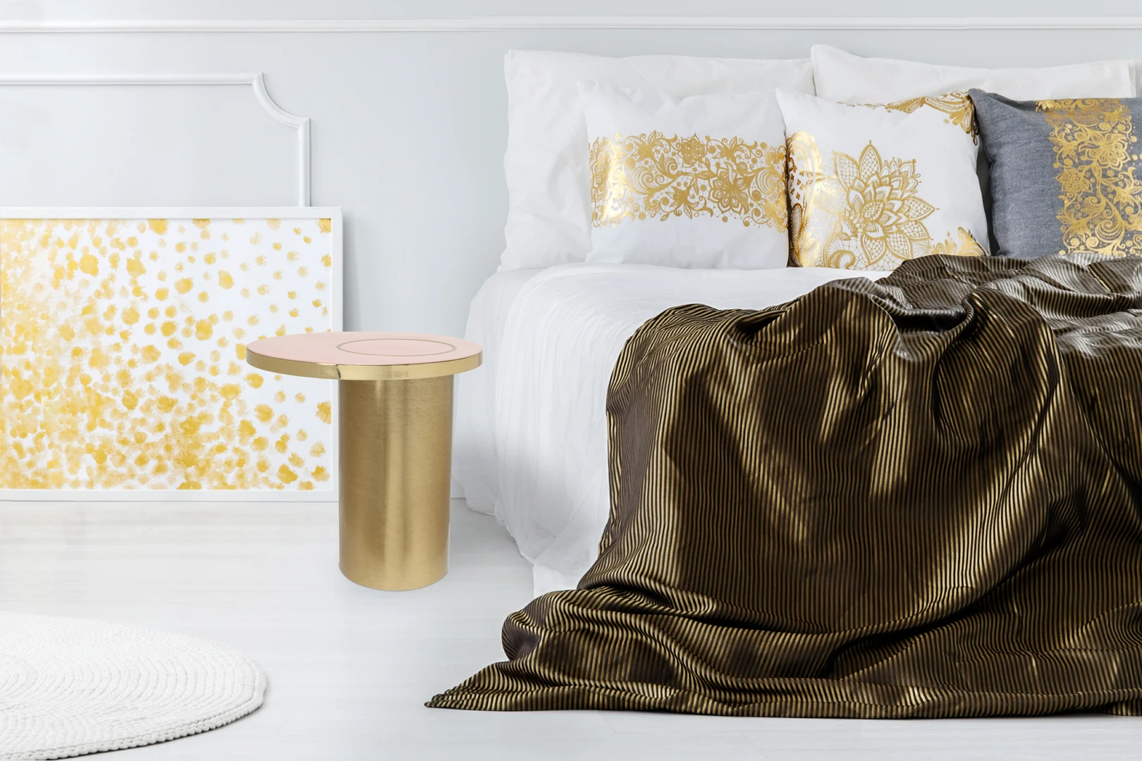 Een moderne slaapkamer met een wit bed, gouden en witte kussens, een goud en bruin gestreepte deken, de Bijzettafel Goud, Roze Rond Ijzer en een wit en gouden abstract kunstwerk tegen de muur.