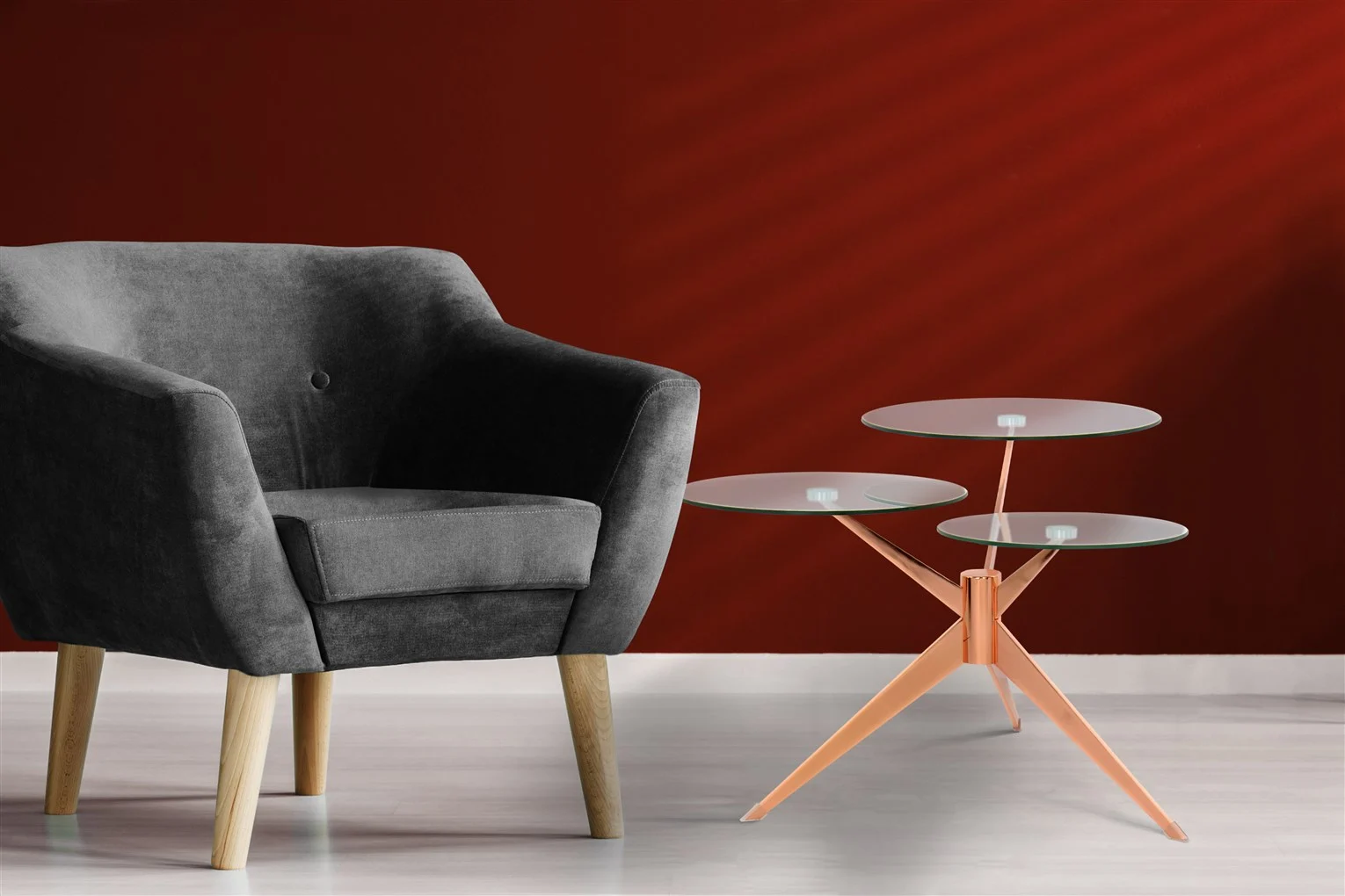 Een moderne grijze fauteuil met houten poten staat naast de Bijzettafel Metaal Compact, een unieke glazen bijzettafel met drie niveaus en een perzikkleurig metalen onderstel, tegen een dieprode muur op een lichte vloer.