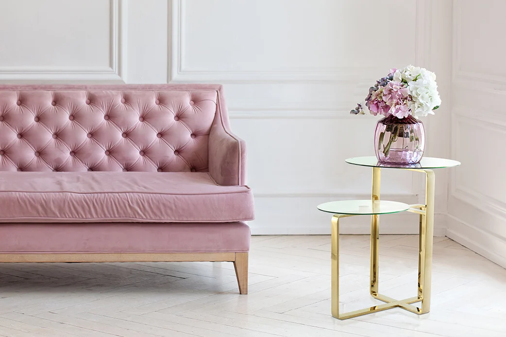 Een roze getufte bank staat naast de Bijzettafel Goud Staal Opbergvak, een bijzettafel van goud en glas. Op de tafel staat een roze vaas met witte en paarse bloemen, tegen muren met witte panelen en een lichte houten vloer.