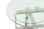 Een close-up van de Bijzettafel Zilver Rond Staal Chique, met een rond glazen blad en glanzend metalen frame met kruisende steunen, die het minimalistische en moderne ontwerp benadrukken.