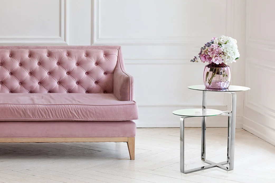 Een roze getufte bank staat naast twee Bijzettafel Zilver Rond Staal Chique tafels, elk bekroond met een glazen vaas met roze en witte bloemen, in een lichte kamer met wit gelambriseerde muren en licht houten vloeren.