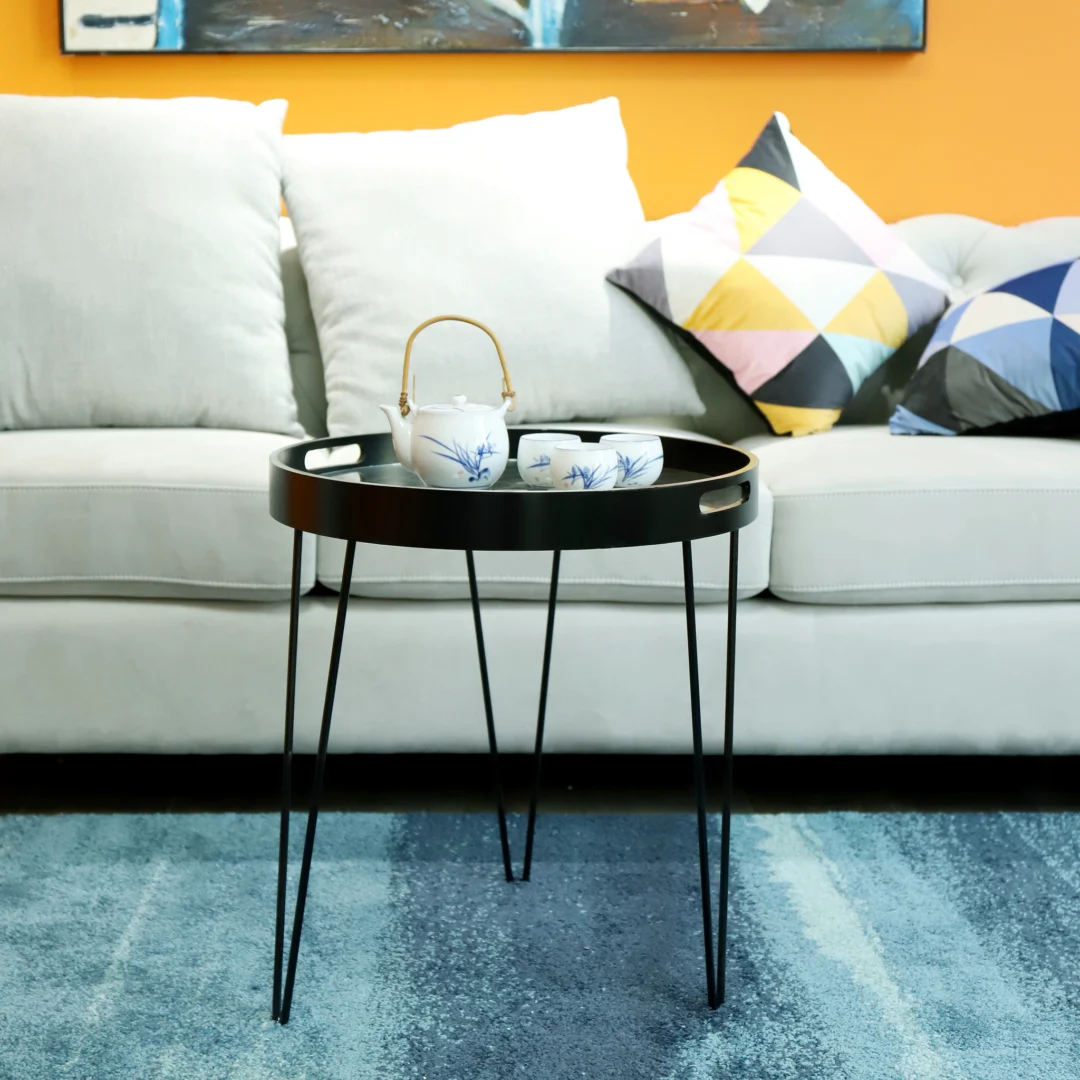 De Bijzettafel Zwart Metaal Industrieel, een rond zwart dienbladtafeltje met een theepot en kopjes, staat op een blauw vloerkleed voor een lichtgrijze bank met gedessineerde en effen kussens tegen een oranje muur.