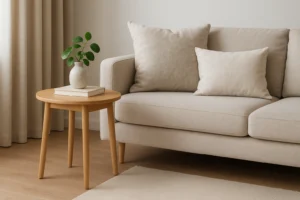 Een beige bank met twee bijpassende kussens staat naast een ronde houten bijzettafel, perfect gekozen met de vraag "wat is de beste hoogte voor een bijzettafel" in gedachten. Natuurlijk licht en een neutrale inrichting creëren een rustige, minimalistische woonkamer.