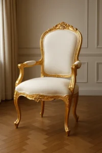 Een elegante fauteuil met een sierlijk gouden frame, crèmekleurige bekleding en uitgesneden bloemendetails staat op een parketvloer in een kamer met wit gelambriseerde muren - perfect in de buurt van een bijzettafel, die zowel in grootte als functie verschilt van een salontafel.