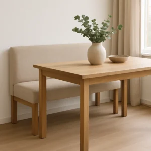 Een minimalistische eethoek heeft een lichte houten tafel - te onderscheiden van een salontafel of bijzettafel - een bijpassende bank met beige bekleding, een witte vaas met groen gebladerte en een ondiepe beige schaal bij een raam met crèmekleurige gordijnen.