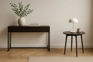 Een minimalistische kamer met een donkerhouten consoletafel - perfect voor wie zich afvraagt "wat is het verschil tussen sidetable en bijzettafel"- heeft een vaas met bladertakken en twee boeken, naast een ronde bijzettafel met een witte lamp en een kopje.