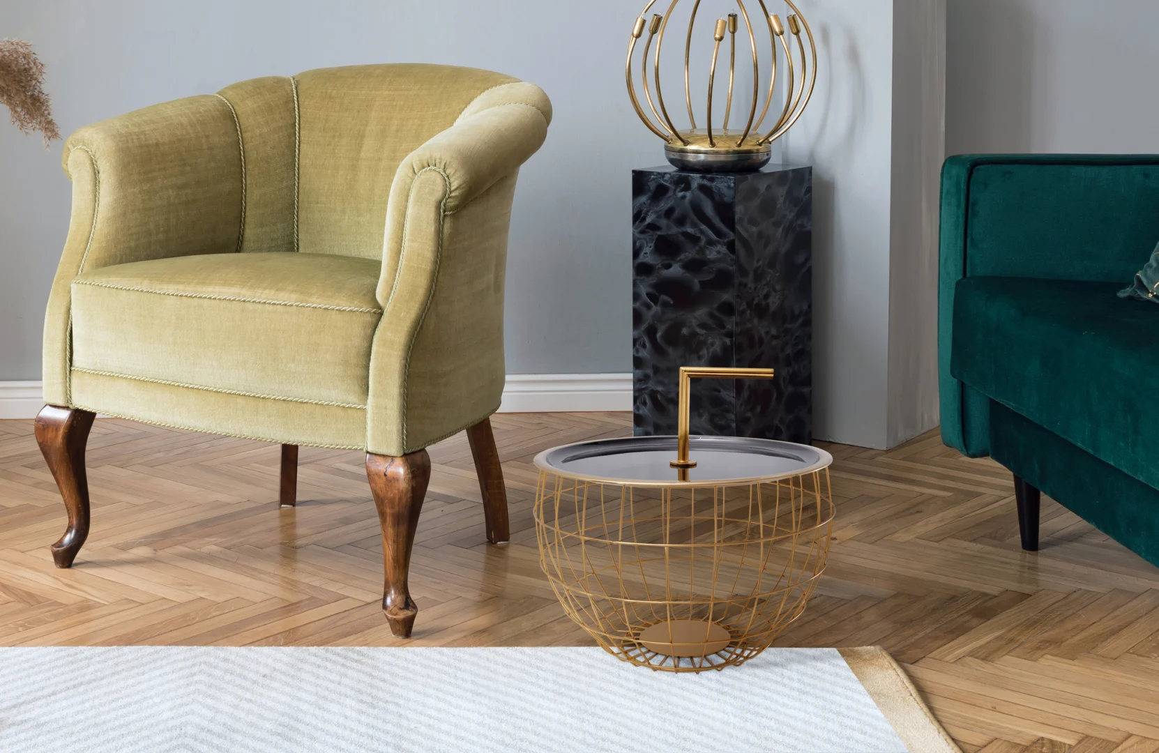 Een stijlvolle woonkamer heeft een fauteuil van groen fluweel, een ronde Bijzettafel Zwart 13 CM Rond Ijzer met opbergruimte waarin een gouden beeldje staat, een salontafel van gouddraad, een groene bank en een houten parketvloer.