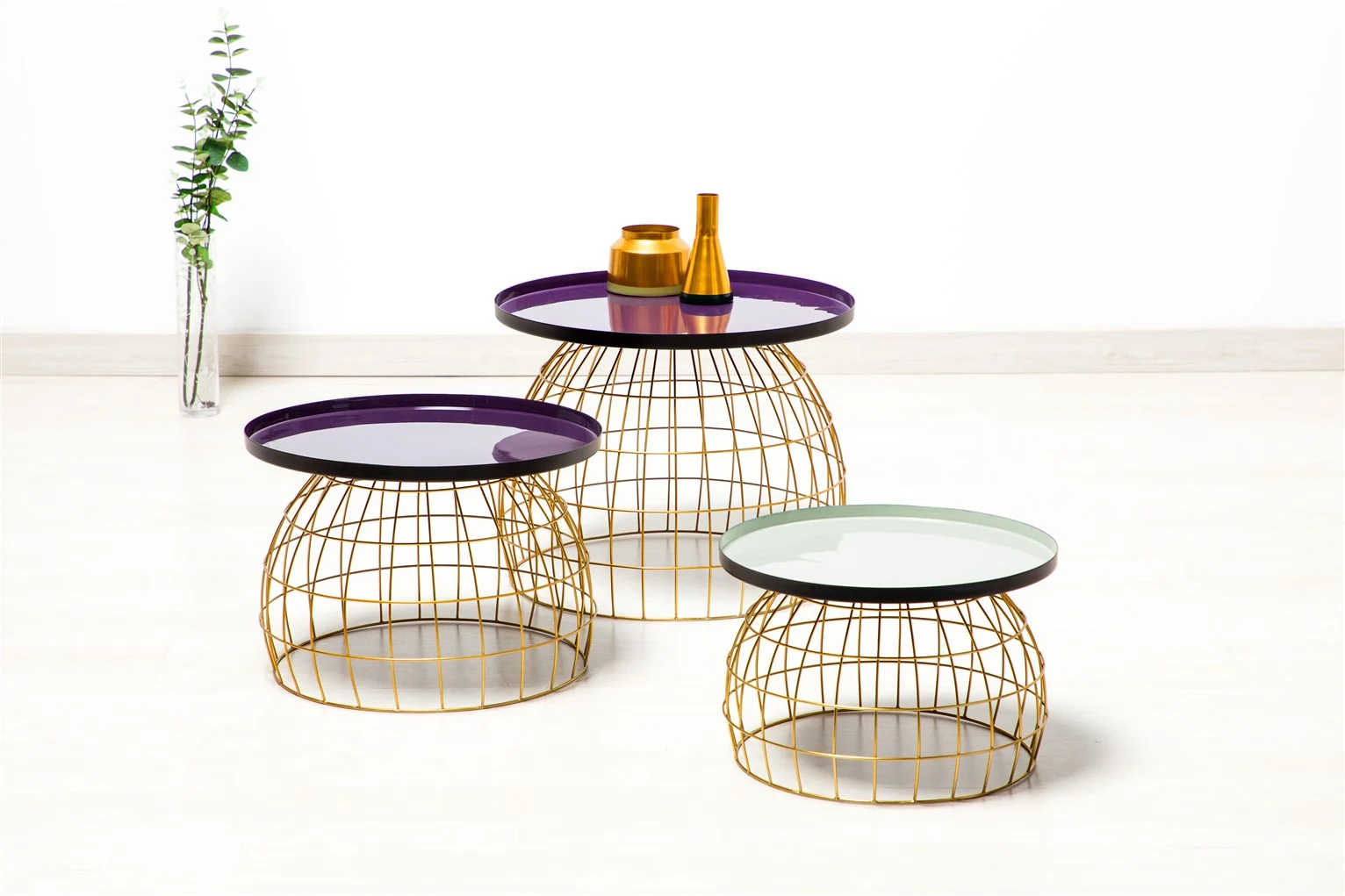 Drie moderne Bijzettafels in lichtgrijs en paarse ronde bladen met onderstellen van gouddraad staan op een lichte vloer, versierd met vazen op de grootste tafel en een hoge vaas met groen op de achtergrond.
