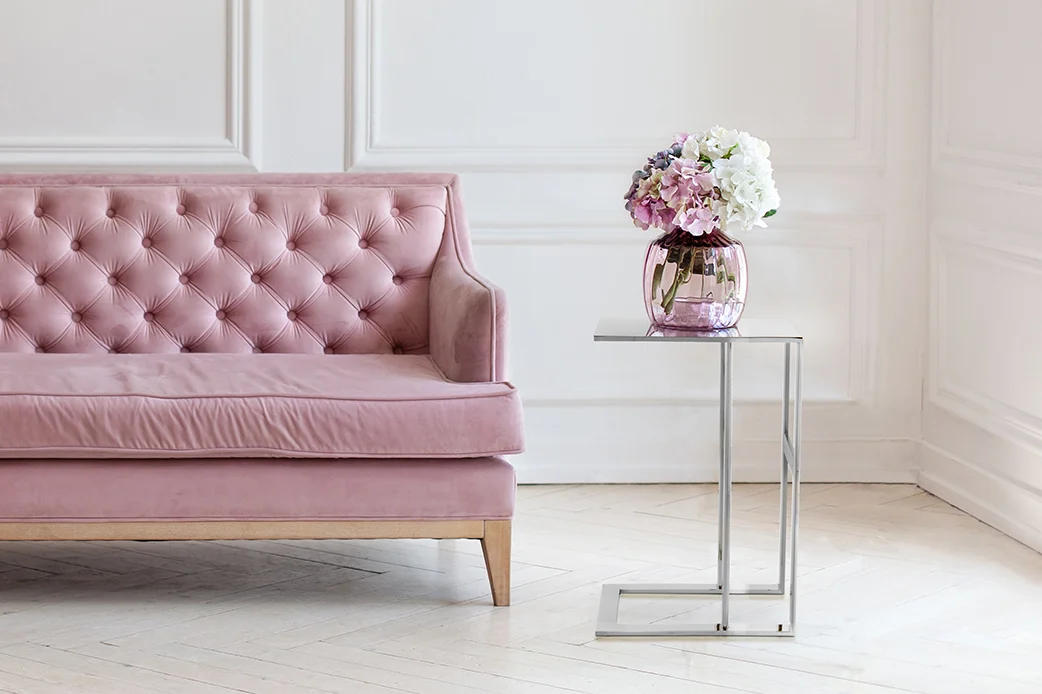 Een roze fluwelen sofa staat naast de Bijzettafel Zilver Rond Staal Compact, een moderne metalen bijzettafel met een glazen vaas met roze en witte bloemen, tegen een wit gelambriseerde muur en een lichthouten vloer.