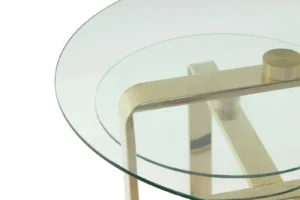 Close-up van de Bijzettafel Goud Staal Design, met een rond glazen tafelblad dat kruist met een hoekig gouden stalen frame, tegen een witte achtergrond.