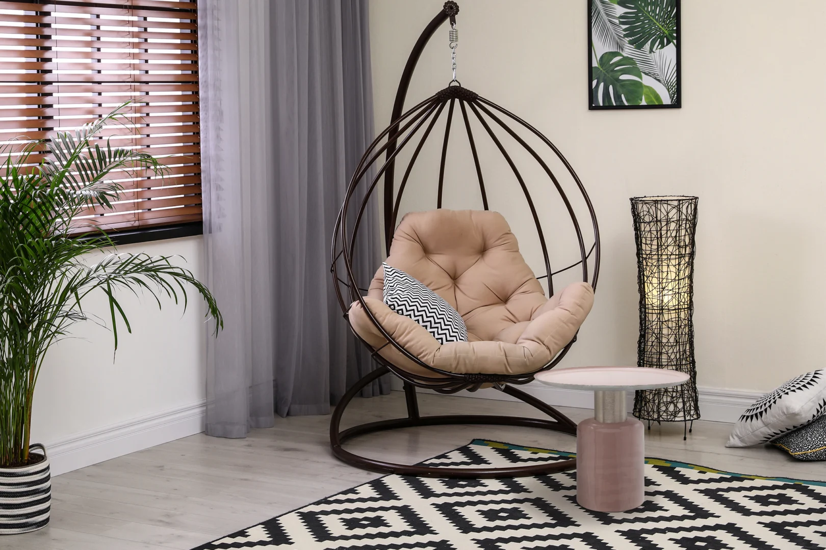 Een moderne woonkamer met een hangende eierstoel, beige kussen, kussen met patroon en de Bijzettafel Zilver Rond Ijzer Chique. Het decor bestaat uit een plant, een decoratieve lamp, een vloerkleed met patroon, gordijnen en muurkunst met bladmotieven.