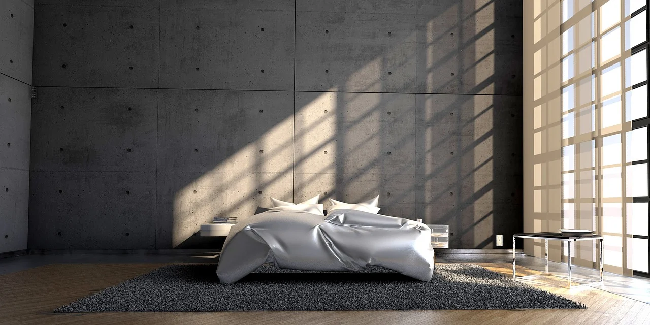 Een moderne minimalistische slaapkamer met een groot onopgemaakt bed op een grijs tapijt, betonnen muren en een raam van vloer tot plafond dat zonlicht en schaduwen door de kamer werpt. Een klein bijzettafeltje en een paar kussens zijn zichtbaar.