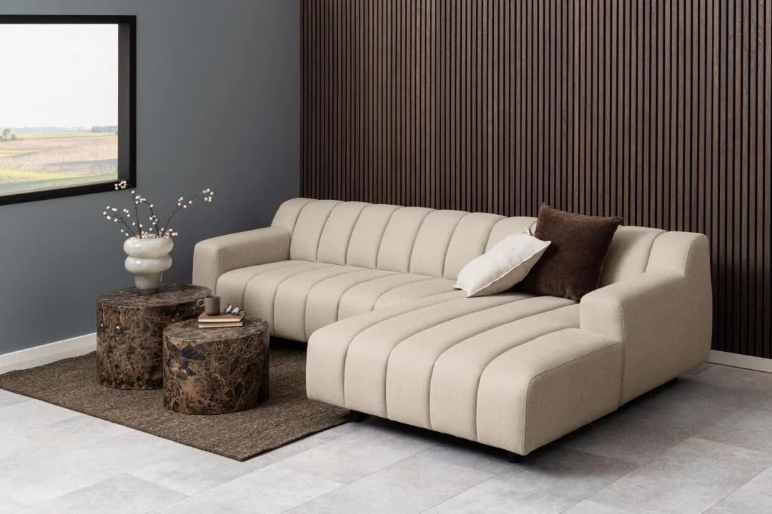 Beige hoekbank met ribbels en bruine kussens. Twee ronde salontafels van donkerbruin kunststof. Zachte belichting, grijze muur en houten panelen.