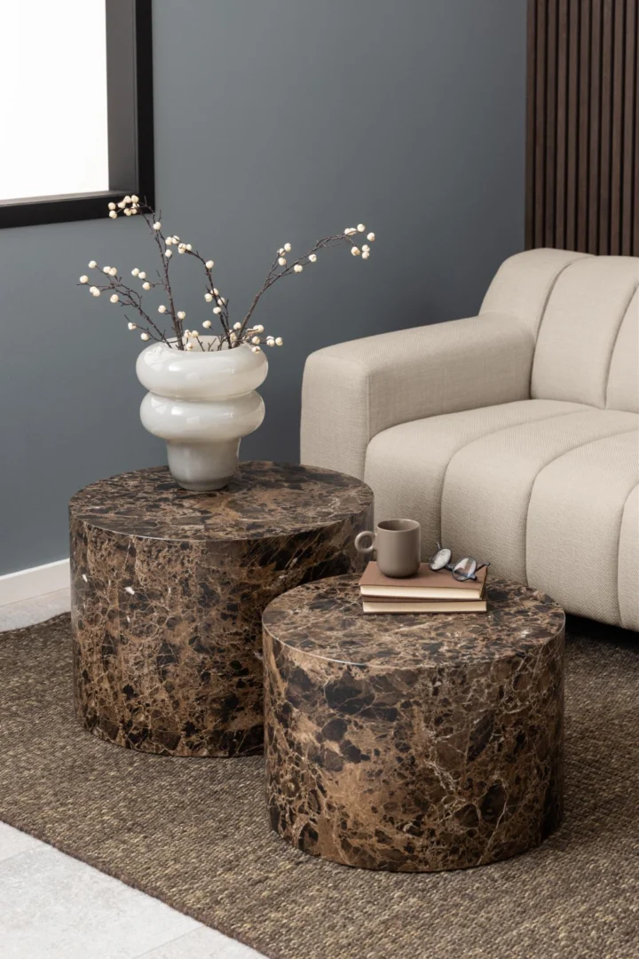 Twee bruine, ronde salontafels van kunststof met marmerprint, een vaas met takken, een kop, boeken en een bril erop, zachte belichting.