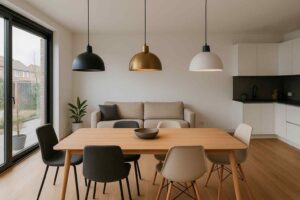 Interieur met houten tafel en stoelen onder drie hanglampen in zwart, goud en wit. Een beige zitbank en plant op de achtergrond. Licht hout.
