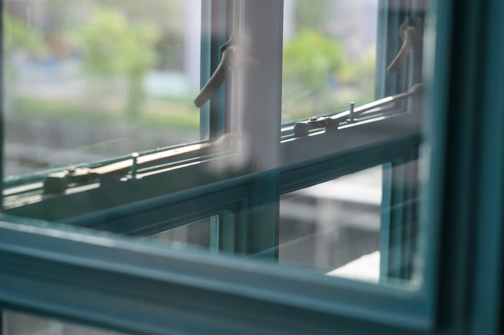 Detailopname van een blauwgroen kozijn met een handgreep en sluiting. Het glas reflecteert de omgeving in zachte, natuurlijke tinten.