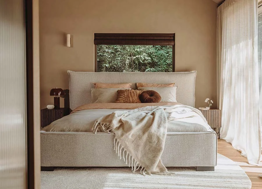 Slaapkamer woonidee: Beige bed met zachte kussens en sprei. Houten nachtkastjes met lampen. Zachte, lichte kleuren en natuurlijk licht.