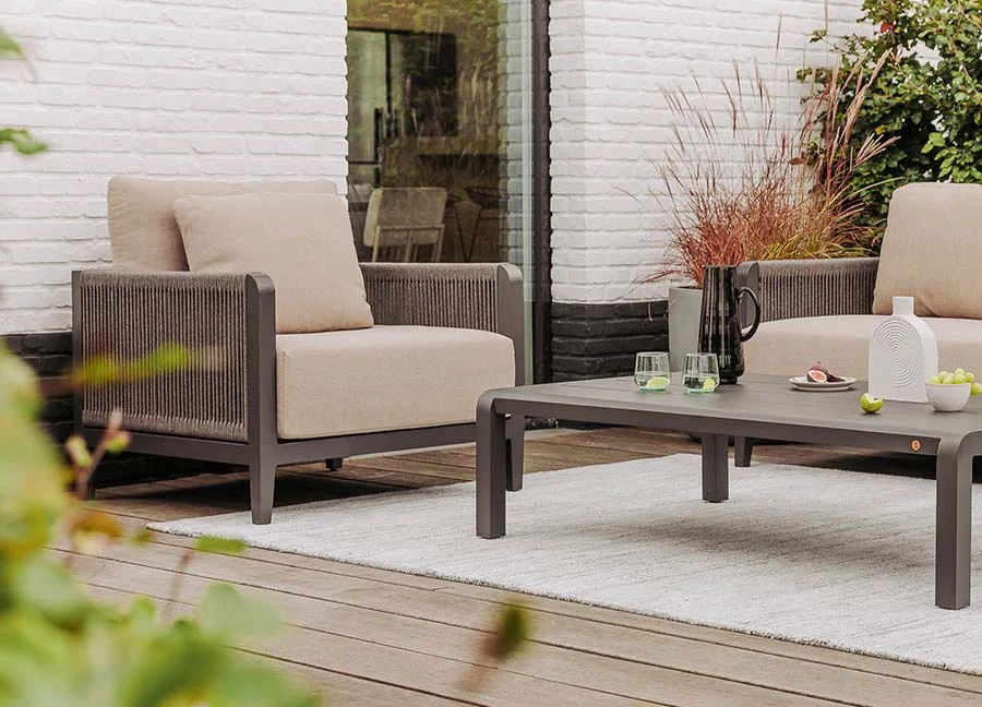 Tuininrichting met beige lounge stoelen en een grijze tafel op een wit vloerkleed. Houten terras en bakstenen muur op de achtergrond.