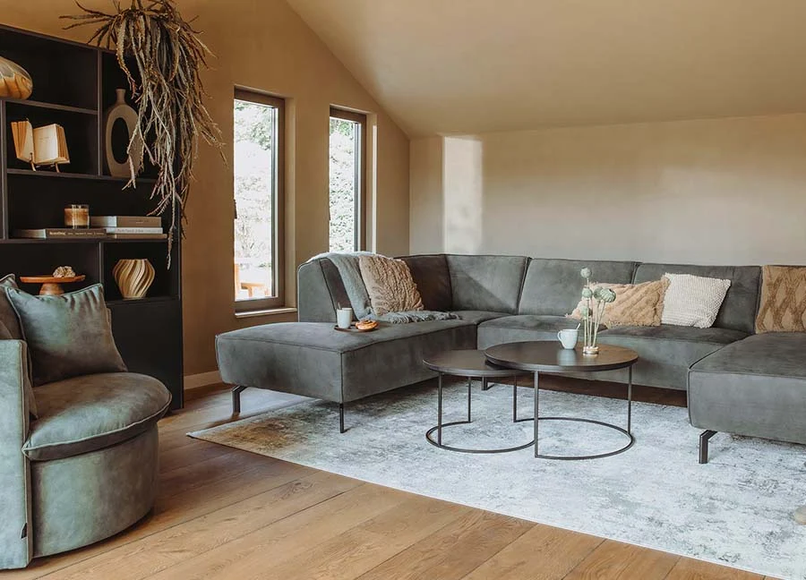 Woonideeën: Hoekbank en ronde tafels in een lichte woonkamer met houten vloer. Zachte kleuren en natuurlijk licht door ramen. Stijlvol interieur.