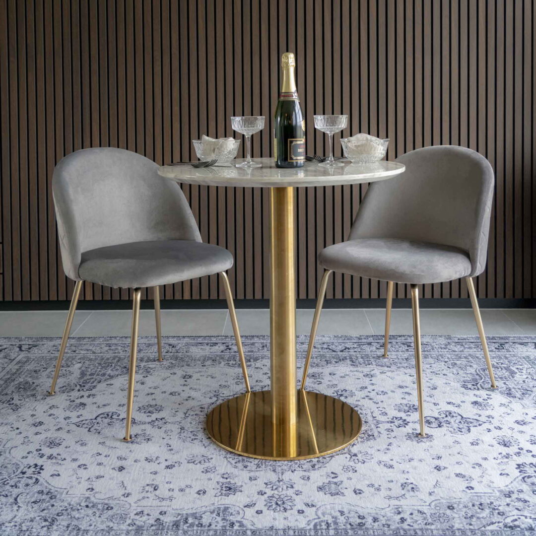 Twee Geneve eetkamerstoelen in grijs velvet met messing poten staan aan een ronde tafel met champagnefles en glazen, op een decoratief tapijt.