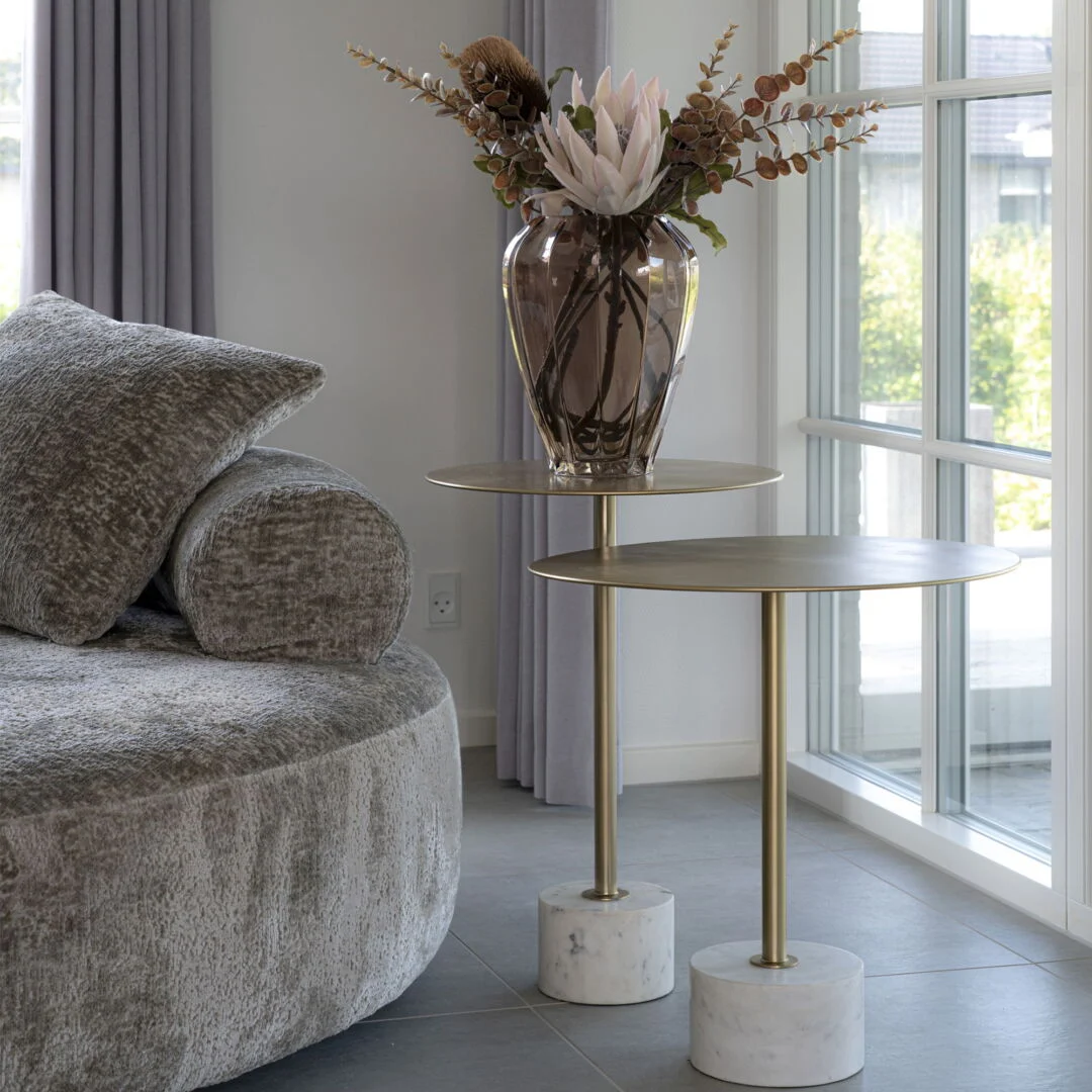 Interior view of a round brass side table with a marble base and a glass vase filled with flowers, naast een grijze bank, in natuurlijk licht.