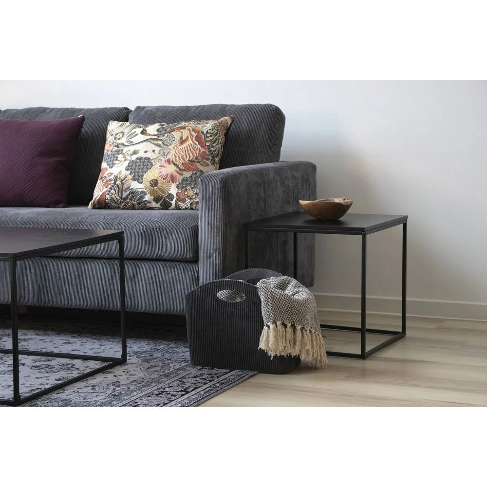 Square black side table with metal frame next to a gray couch with patterned pillows, styled with a blanket and magazine holder in neutrale kleuren.