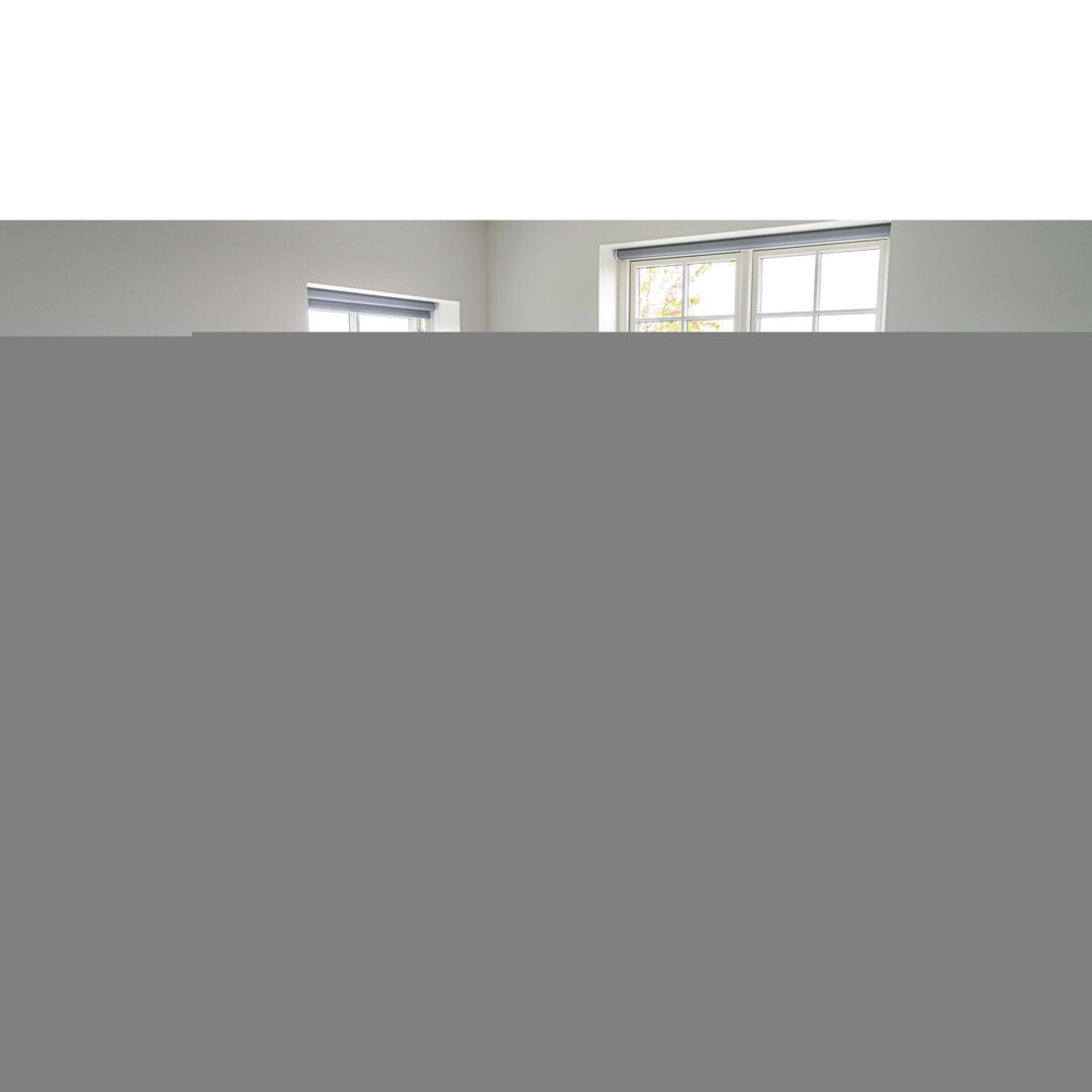 Lage hoekopname van een lege kamer met twee ramen met witte kozijnen en grijze rolgordijnen tegen een lichte, zachte achtergrond van buiten.