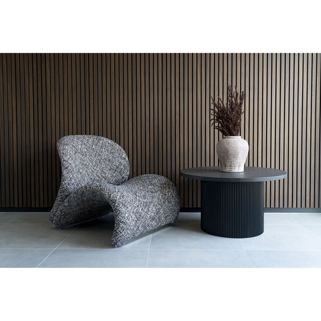 Stylish interior featuring a grijs gemêleerde designstoel naast een ronde salontafel met een zwarte geribbelde basis tegen een houten lambrisering.