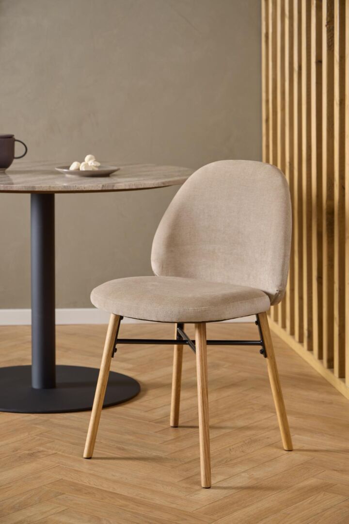 Sophia eetkamerstoel: Beige stoffen stoel met houten poten naast een ronde tafel.