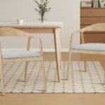 Eetkamerstoel Claire Essenhout Naturel Beige: Twee houten stoelen met beige kussens bij een tafel.
