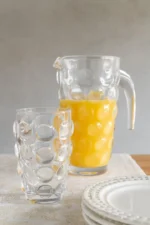 Transparant Limonadeglas Ciessino van glas met reliëf, naast karaf met sinaasappelsap op gedekte tafel