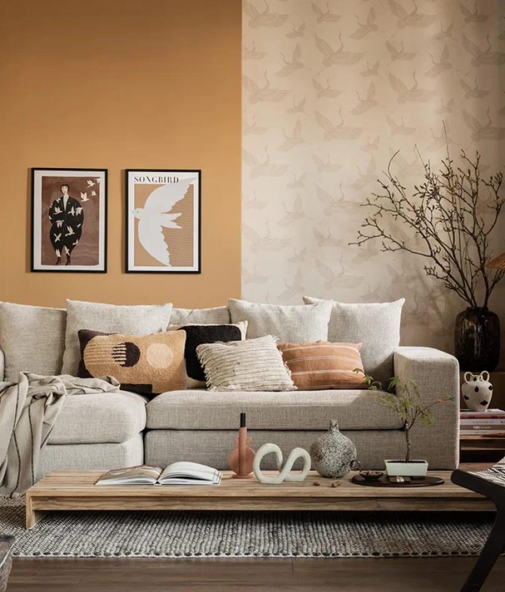 Een gezellige woonkamer heeft een beige sofa met verschillende neutrale kussens, een houten salontafel met decor, muurkunst en muren met warme tinten. Natuurlijk licht valt binnen door een raam, waardoor een aardse en minimalistische inrichting wordt benadrukt.