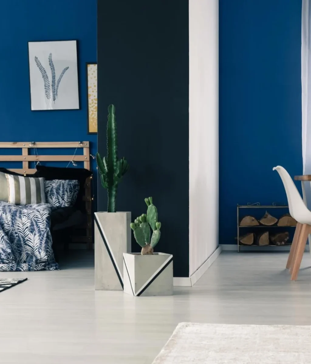 Moderne woon- en eetkamer met felblauwe muren, gedessineerde kussens, een houten lamp, cactussen in geometrische potten, witte eetkamerstoelen en natuurlijk licht dat door gedessineerde gordijnen naar binnen valt.