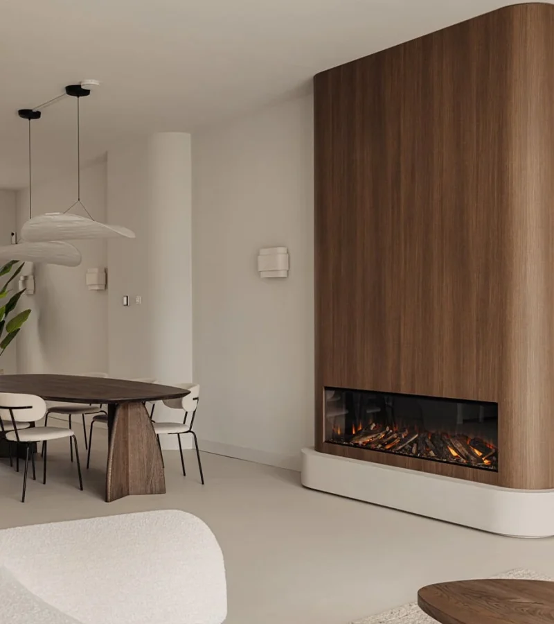 Een moderne eet- en woonruimte met neutrale tinten, met een houten eettafel met witte stoelen, een grote plant, minimalistische hanglampen en een ingebouwde elektrische open haard met een hoge houten rand.