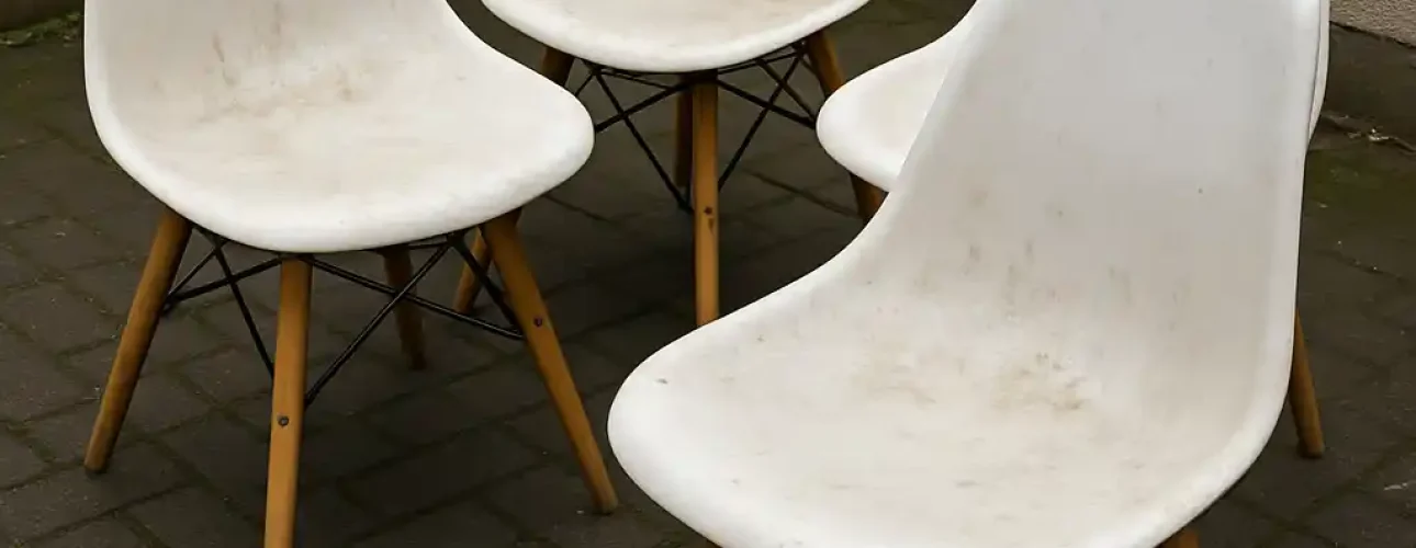 Vier witte plastic stoelen met houten poten staan buiten op een bakstenen patio bij een bakstenen muur en een witte muur. De stoelen lijken wat vies-krijg je witte stoelen weer stralend schoon met een goede reiniging.