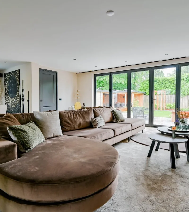 Een ruime, moderne woonkamer met een grote bruine sofa, ronde salontafels en glazen deuren van vloer tot plafond die openen naar een groene achtertuin. De eethoek en decoratieve planten zijn zichtbaar op de achtergrond.