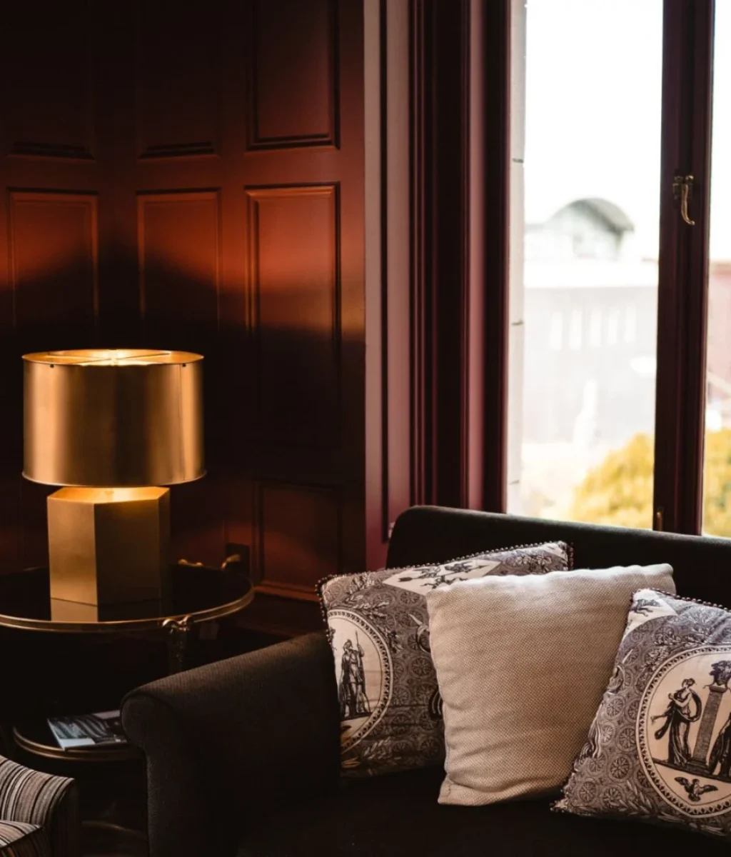 Een gezellige woonkamer met een donkere bank en kussens met patroon, een fauteuil, een bijzettafel met een gouden lamp en grote ramen die natuurlijk licht binnenlaten met uitzicht op de stad buiten.