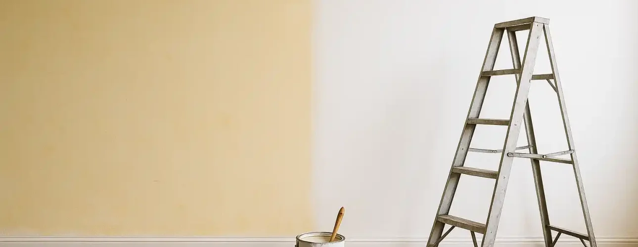 Een trapladder staat naast een muur die gedeeltelijk van beige naar wit is geschilderd om te bewijzen dat je vergeelde verf weer wit kunt krijgen. Een verfblik, kwast en hamer rusten op een droogdoek die de hardhouten vloer bedekt.