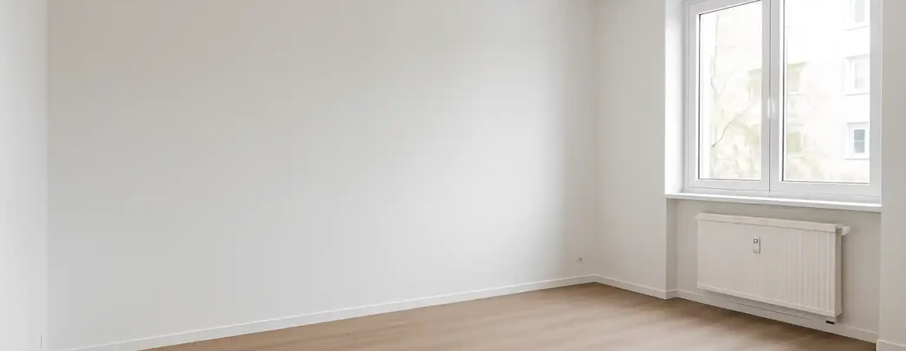 Een lege kamer met witte muren -dat gevoel krijg je van witte muren- heeft een groot raam dat natuurlijk licht binnenlaat, een witte radiator eronder en lichtgekleurde houten vloeren.