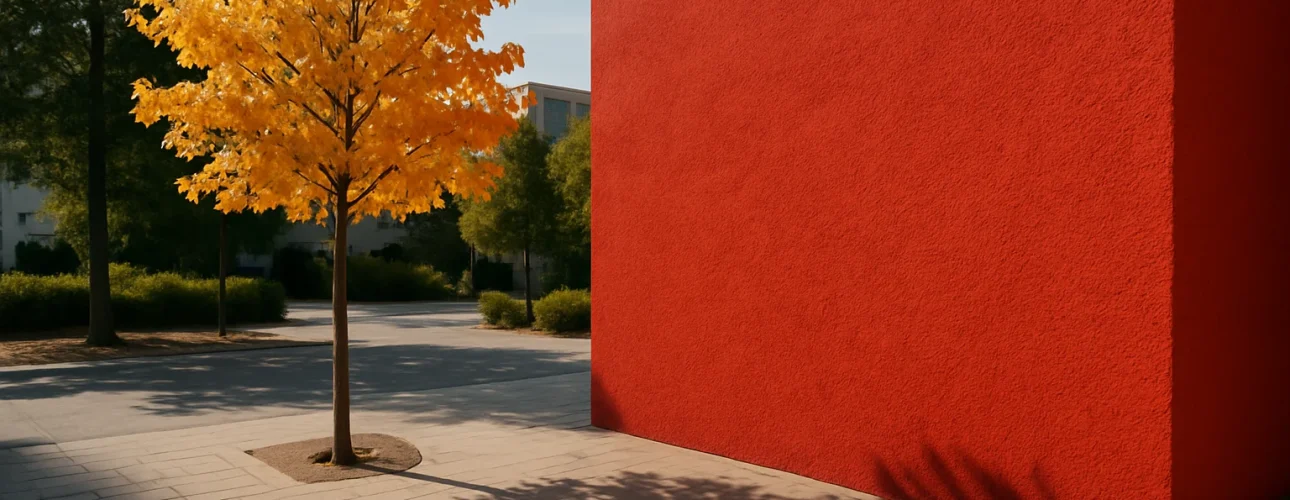 Een kleine boom met felgele bladeren staat naast een grote, levendige rode muur - een tafereel waarvan je je afvraagt: welke kleur is mode? Zonlicht werpt heldere schaduwen op de geplaveide binnenplaats, groene bomen zorgen voor een levendig contrast op de achtergrond.