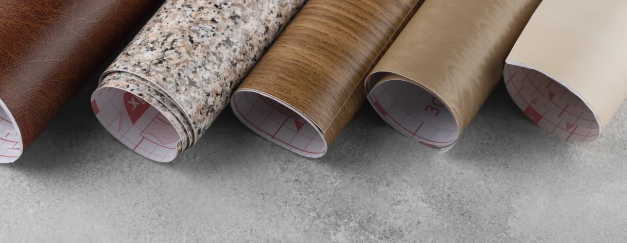 Vijf rollen zelfklevend contactpapier met verschillende patronen - bruin leer, graniet, houtnerf, lichtbruin en beige - worden naast elkaar gelegd op een grijs betonnen oppervlak.