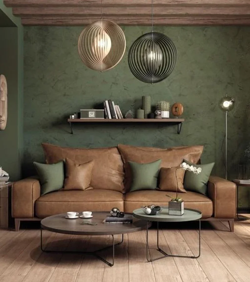 Gezellige woonkamer met een bruine leren bank, groene accentkussens, ronde salontafels, hangende hanglampen, een houten fauteuil, planten en een groene muur met structuur, waardoor een warme en uitnodigende sfeer ontstaat.